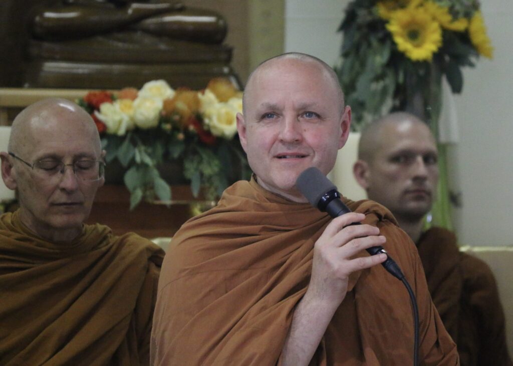 Three Thai Forest Theravada Buddhist monks in ocher robes sit on a stage in front of flowers on an altar. One is speaking into a microphone.